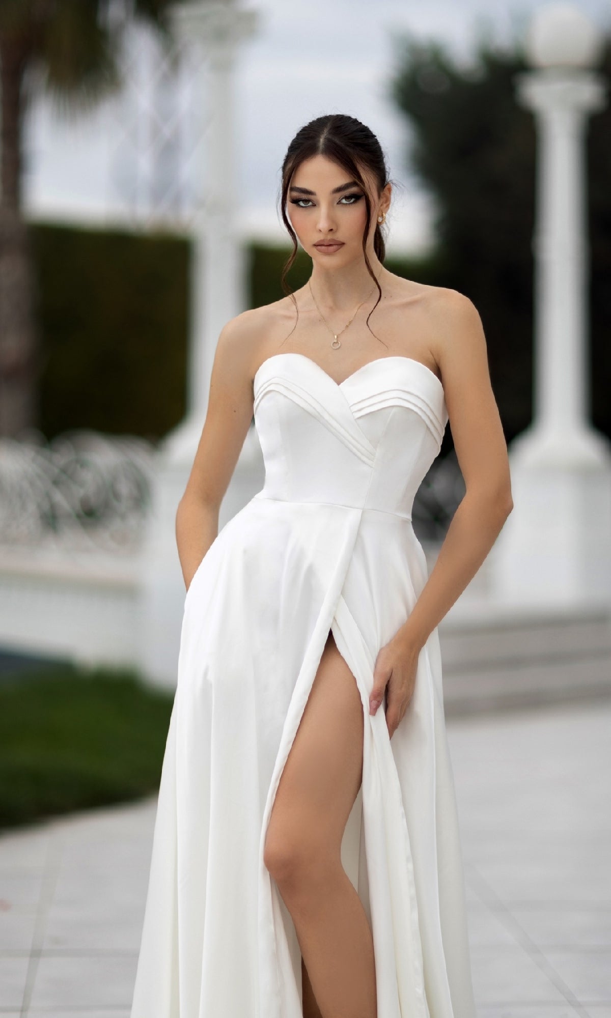 A woman stands outdoors on a tiled surface wearing the Off-White Strapless Bridal Gown: Tina Holly TW105, featuring a sweetheart neckline and a thigh-high slit.